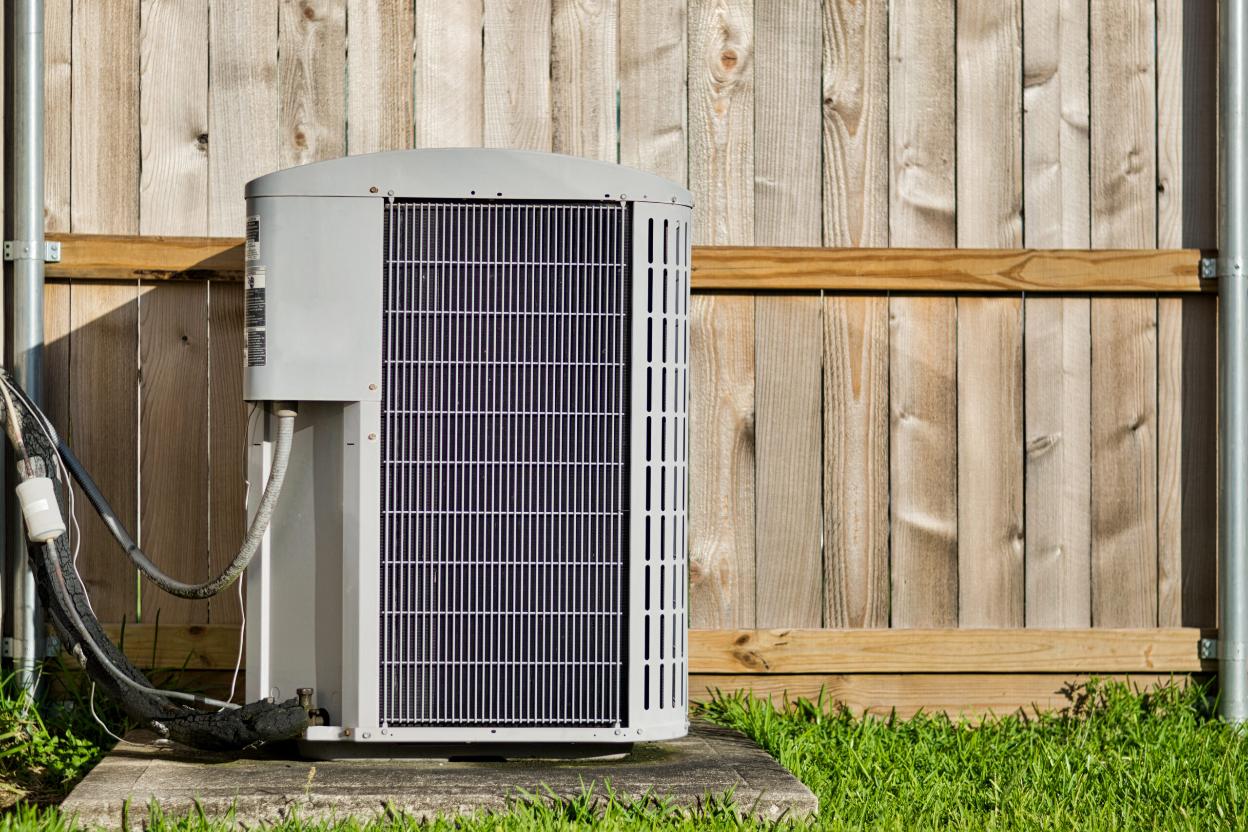 Central air conditioning unit in a residential backyard in front of a defocused wooden fence and lawn area with copy space.